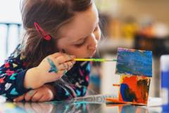 A three year old girl paints a tiny birdhouse with water colour paints.